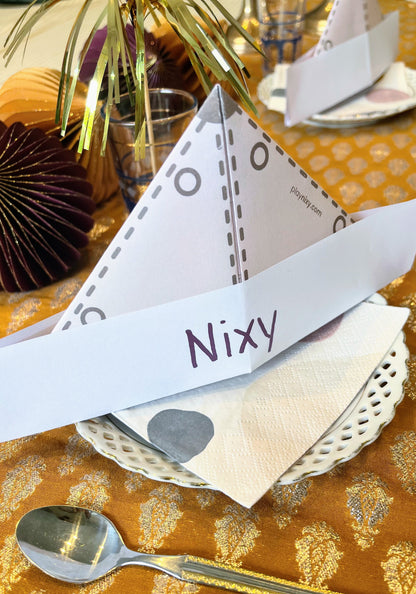 Safari hat place card with 'Nixy' on a decorative table setting with gold tablecloth and silver spoon.